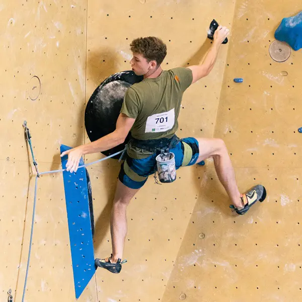 Een klimmer met een klimgordel en nummer 701 beklimt een indoor-klimwand, waarbij hij zich vasthoudt aan een zwarte en een blauwe greep terwijl hij zich opmaakt voor de volgende beweging.